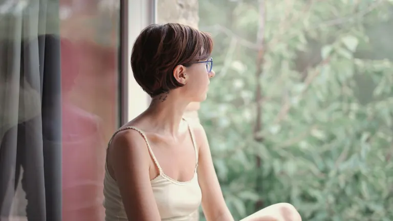 A woman sitting by a window, looking out at green trees